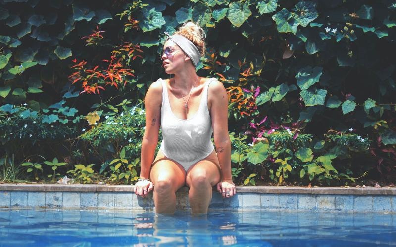 woman in bathing suit and sunglasses sits on pool ledge with her legs in the water